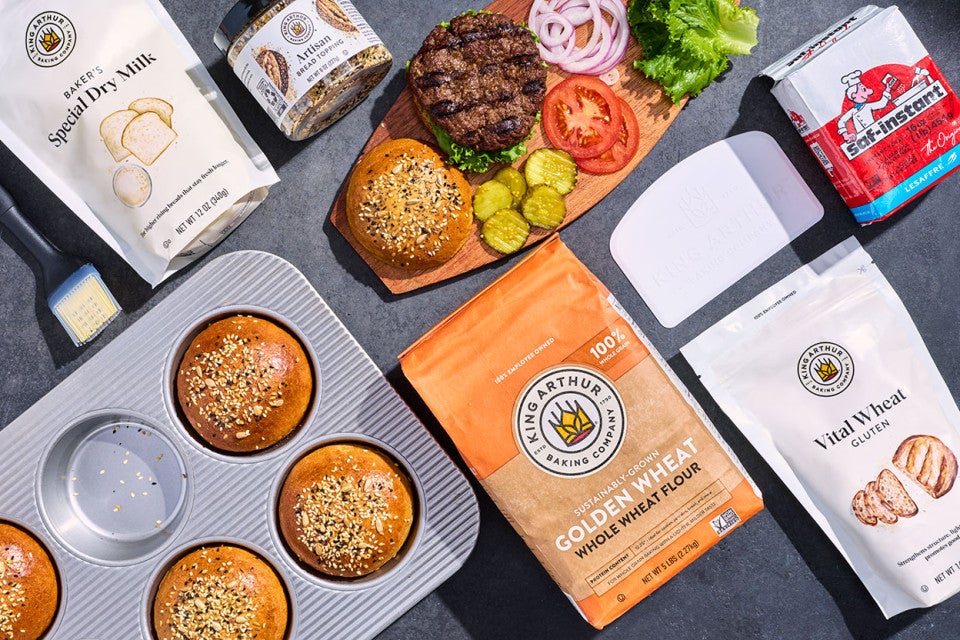 Whole Wheat Hamburger Buns in a bun pan with the ingredients to make them arranged on a counter.
