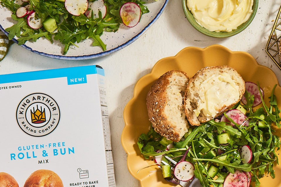 A Gluten-Free Seeded Hearth Roll on a plate with a box of Gluten-Free Roll & Bun mix.