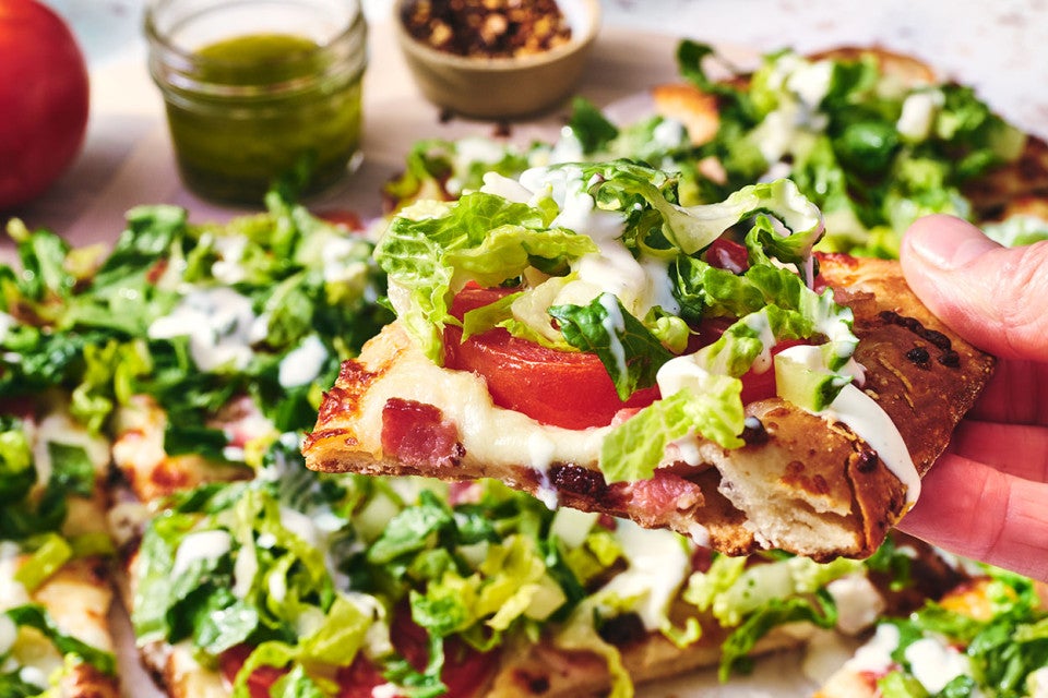 A slice of BLT Pizza shown from the size with tomatoes, bacon, and fresh chopped lettuce