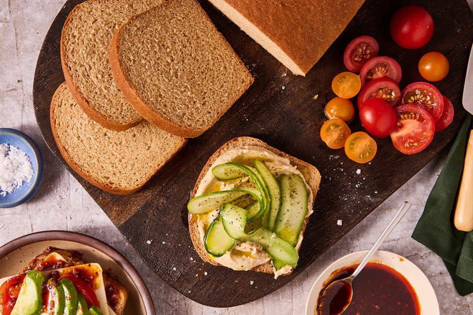 Classic 100% Whole Wheat Bread sliced on a cutting board with tomatoes, cucumbers, and other elements.