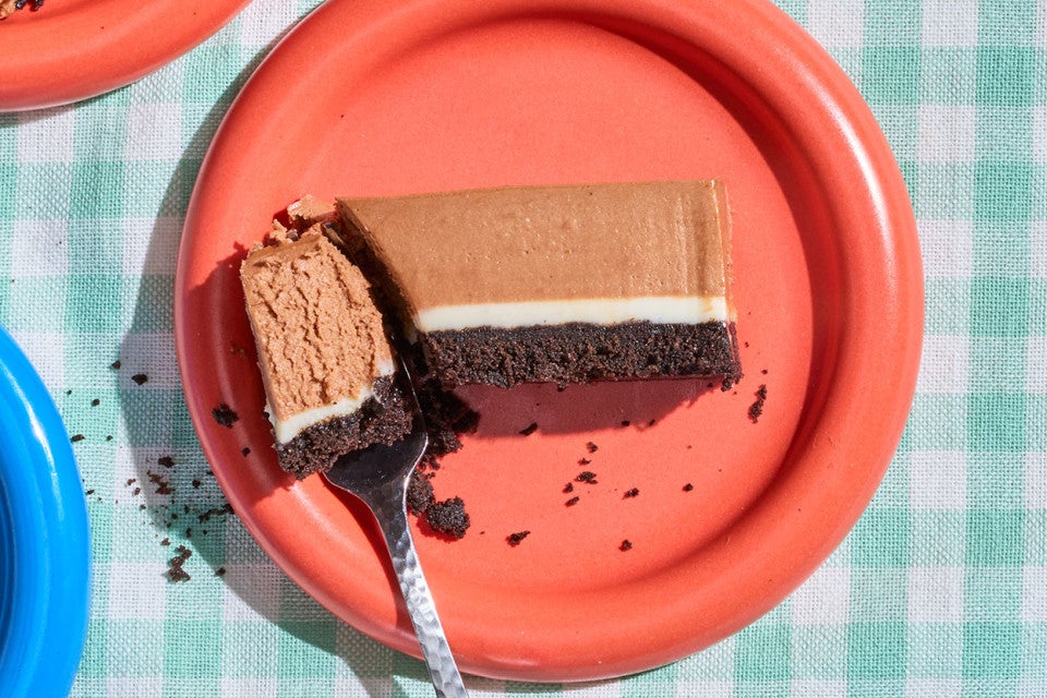 A bite of a No-Bake Chocolate Cheesecake Bar on a fork seen from overhead with a red plate and a teal checkered tablecloth
