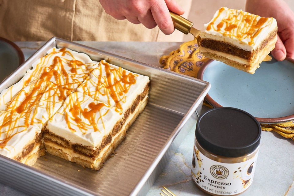 Putting a slice of Caramel Macchiato Tiramisu on a plate with the rest of the baked good in a baking sheet and a jar of King Arthur Baking Espresso Powder in the foreground.