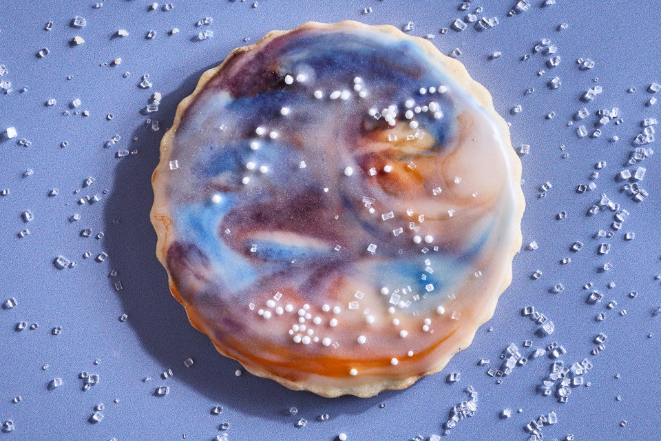 A Northern Lights Iced Sugar Cookie on a blue background surrounded by sprinkles of sparkling sugar