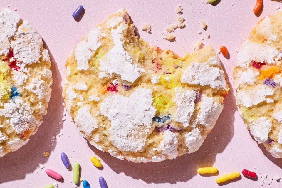Sprinkle Crinkle Cookies on a light lavender background and a bite taken out of a cookie