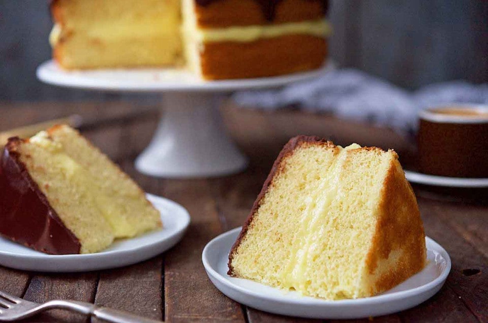 A Boston Cream Pie on a cake stand featuring two layers of yellow cake filled with pastry cream and a chocolate ganache, with two slices on plates in front of it.