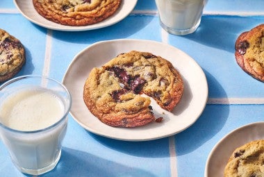 A Classic Chocolate Chip Cookie split on a plate atop a blue counter with glasses of milk in the photo.